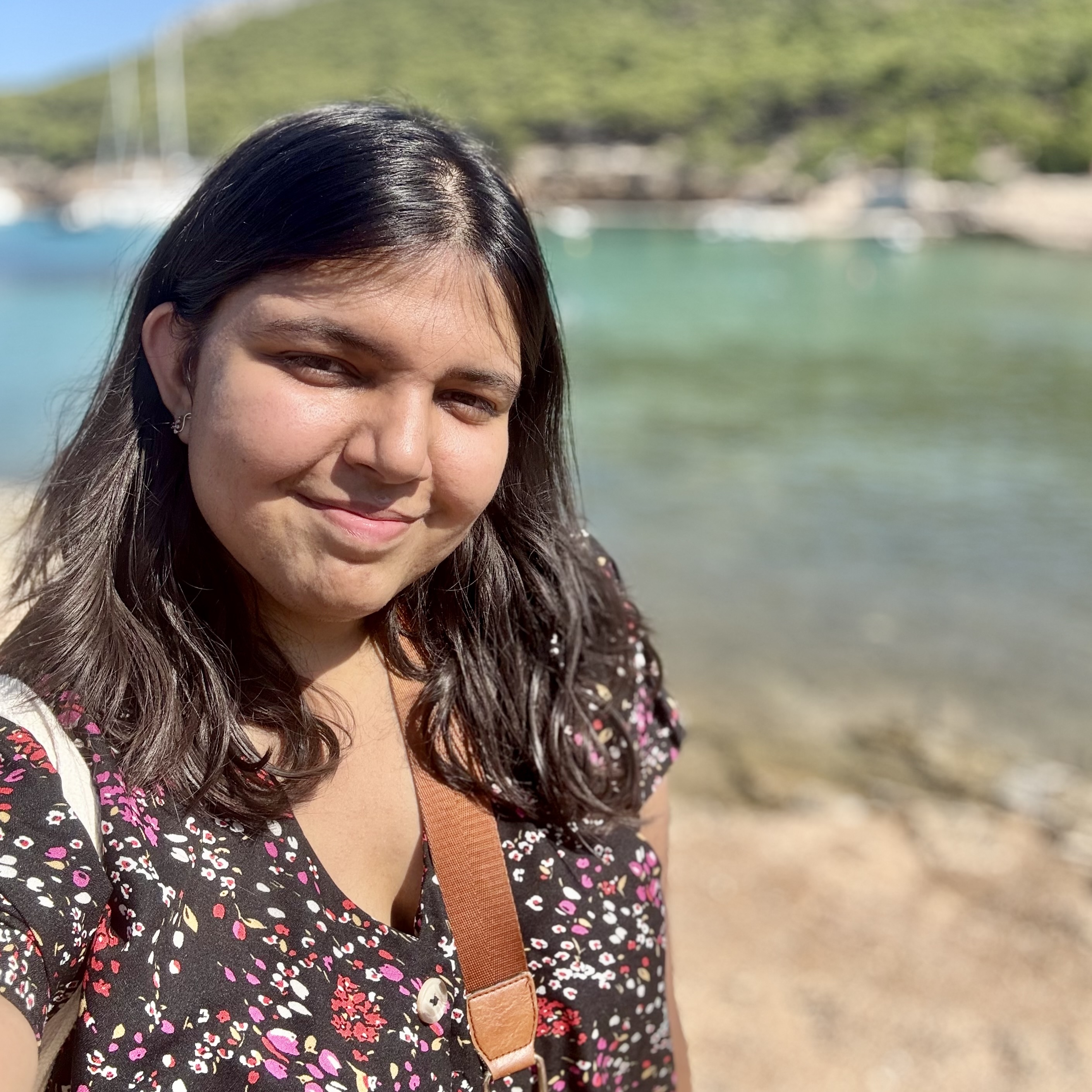 An Indian girl, wearing a black floral shirt, standing in front of aqua blue water and trees, squinting in the sunlight.